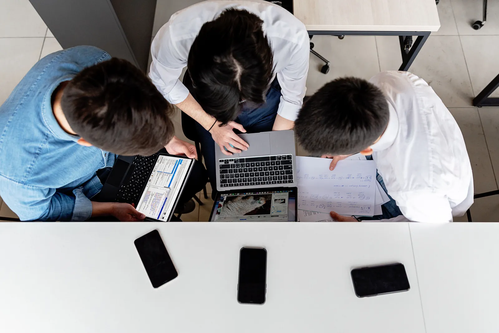 Trei persoane colaborează la laptopuri, cu documente și telefoane pe masă