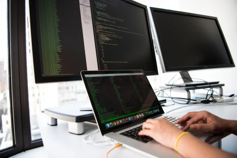 O persoană scrie cod pe un laptop lângă monitoare